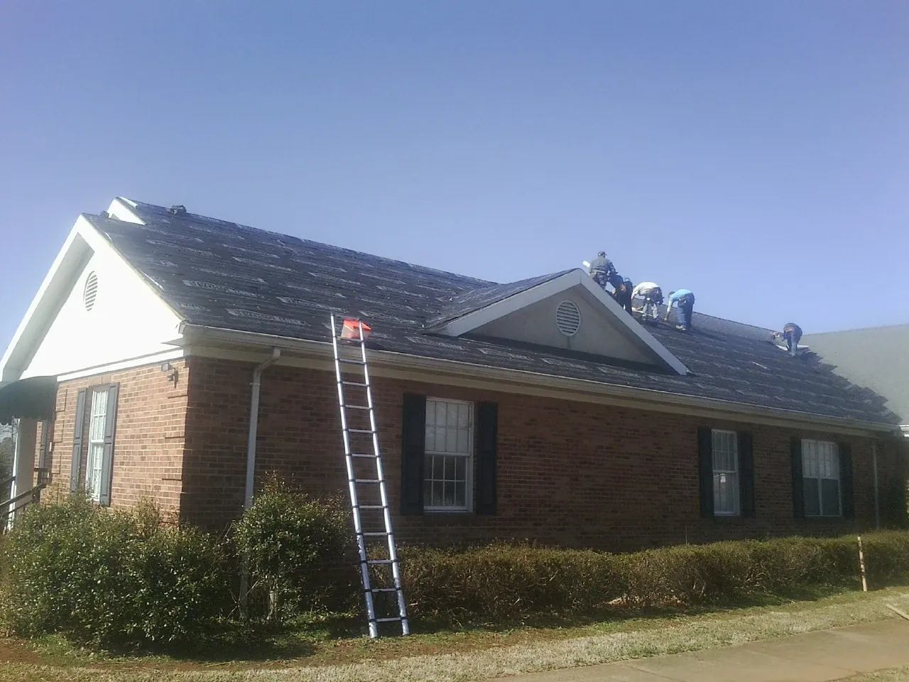 Crew tearing shingles off a brick ranch roof with a ladder set — Emergency Roof Repair work by our crew in Carencro