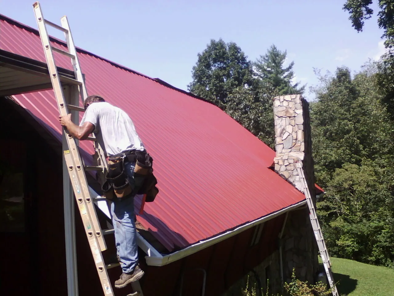 Roofer on a ladder beside a red metal roof and stone chimney — Soffit & Fascia Repair by Roofing Brothers in Carencro